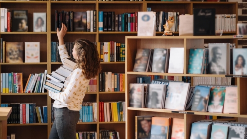 Beautiful girl is holding stack of books while standing among books in the bookshop