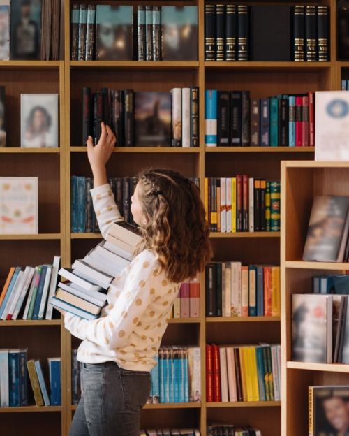 Beautiful girl is holding stack of books while standing among books in the bookshop