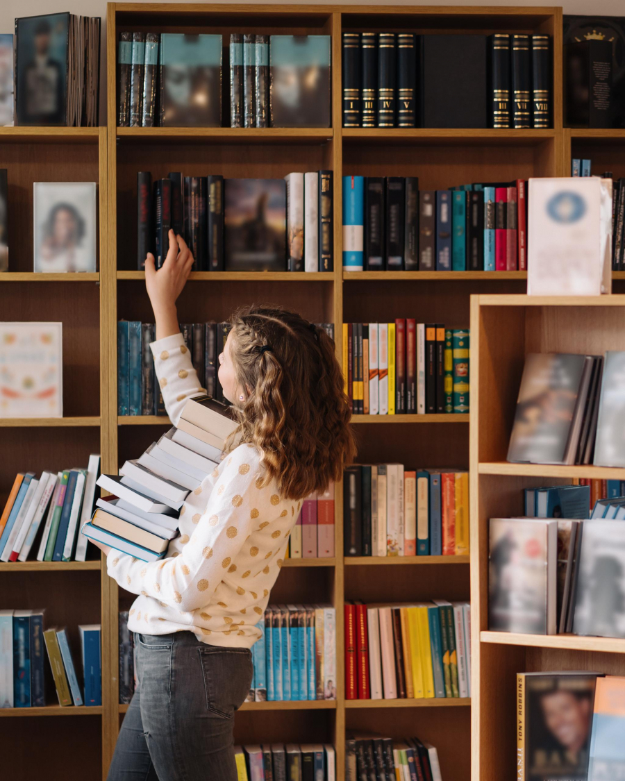 Beautiful girl is holding stack of books while standing among books in the bookshop